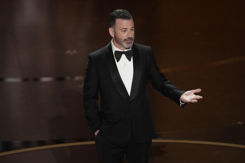 Jimmy Kimmel in a black tuxedo and bow-tie standing and speaking on a stage with his right hand out