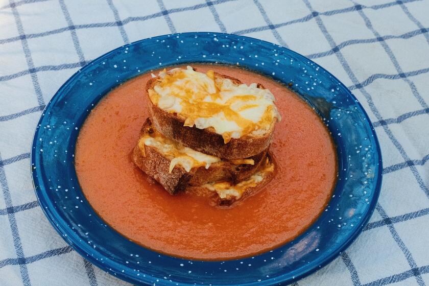 A blue enamel bowl of tomato soup on white-and-blue checkered cloth. At center of bowl is a stack of cheese-topped crostini