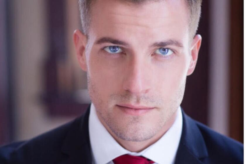 Actor Paul Teal appears in a head shot wearing a dark blazer, white shirt and red tie