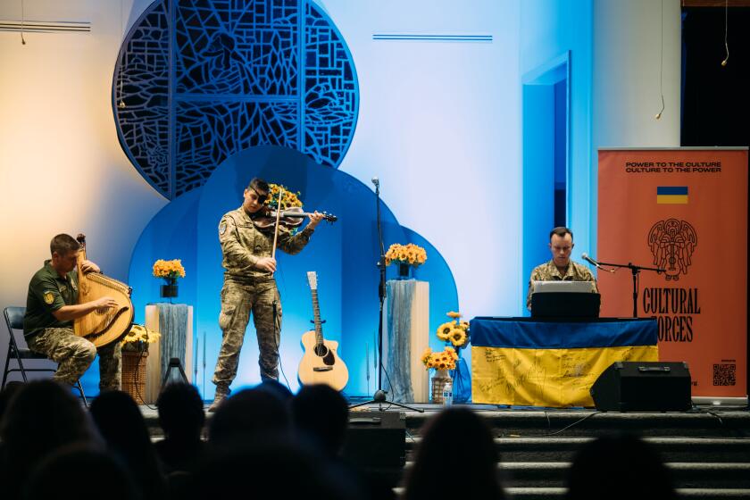 A bandura player, violinist and keyboard player perform in what appears to be a church, with the Ukrainian flag displayed. 