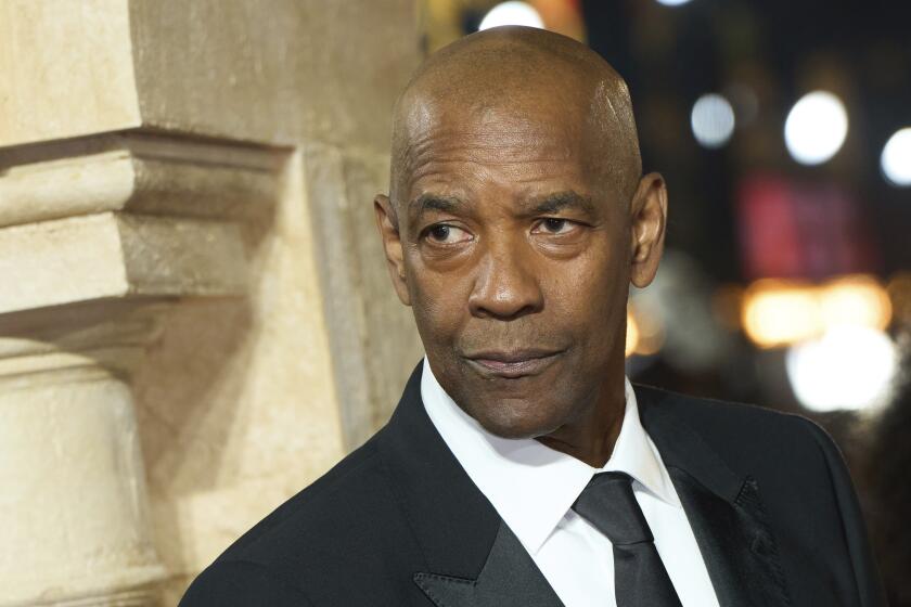 Denzel Washington in a black suit and tie leaning forward and posing against a column