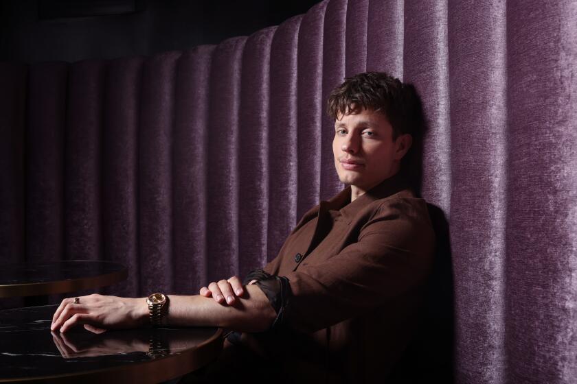 Matt Rife in a brown suit sitting up against a purple velvet wall and resting his left arm on a dark table