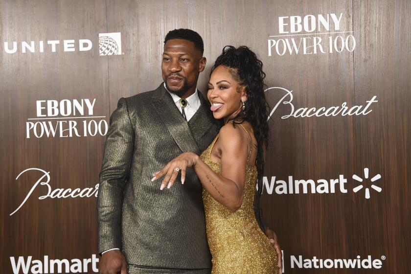 Jonathan Majors in a metallic green suit and tie posing next to Meagan Good in a gold dress against a brown background