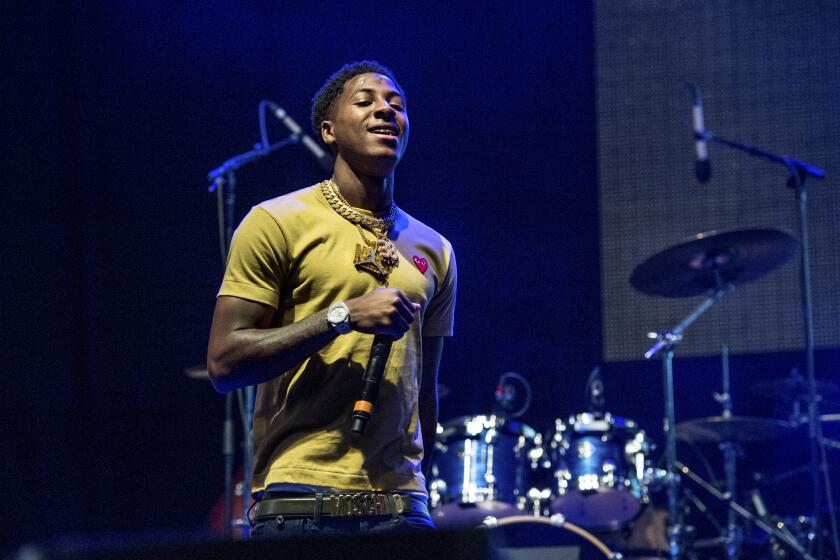 Rapper NBA YoungBoy in a mustard yellow shirt on a stage holding a microphone with his right hand